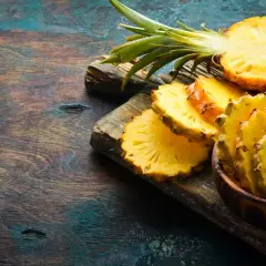 Frische Ananas-Scheiben, eine halbe Ananas und eine ganze Frucht liegen auf einem rustikalen Holzbrett. Das Fruchtfleisch ist leuchtend gelb und zeigt die typische Struktur, die auch für Bromelain bekannt ist.