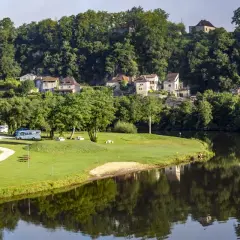 Vue paisible d'un camping-car près de la rivière, avec des maisons pittoresques accrochées à la colline, rappelant le charme de Figeac et Cahors.