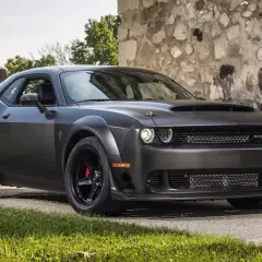 Mroczny Dodge Challenger Demon, z potężną maską i agresywnym grillem, stoi przed kamienną ścianą.
