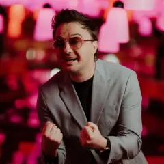 Mężczyzna w okularach i garniturze, z zaciśniętymi pięściami, świętuje sukces. W tle różowe lampy, idealne na after party wiek.