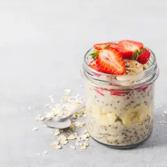 En burk med overnight oats recept toppad med jordgubbar och banan. Havregryn och en sked ligger bredvid.