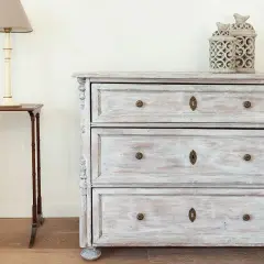 Un meuble cérusé patiné, avec ses tiroirs et ses ornements délicats, se dresse à côté d'une petite table d'appoint surmontée d'une lampe.