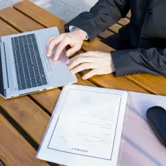 Biznesmen przy laptopie analizuje, ile kosztuje założenie księgi wieczystej. Na stole dokumenty i etui na okulary.