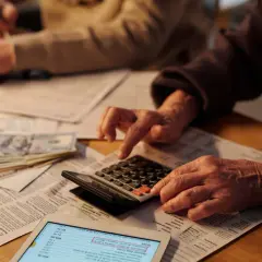 Osoby liczą na kalkulatorze dokumenty, wśród których leżą pieniądze. Prawdopodobnie chodzi o podatek od spadków i darowizn.