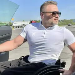 Mężczyzna na wózku inwalidzkim przy otwartych drzwiach samochodu na parkingu.