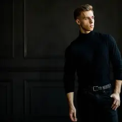 Młody mężczyzna w czarnym golfie i jeansach, pozujący na tle ciemnej ściany.
