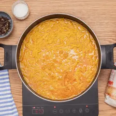 Fideuá con almejas burbujeando en una sartén. Alrededor, especias, perejil y un paquete de fideos.