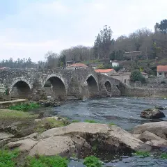 El ponte Maceira, un puente de piedra con arcos, cruza un río caudaloso. Casas con tejados rojos se asoman en la ladera.