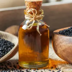 Naturalny olejek z czarnuszki na twarz w szklanej butelce, obok drewnianych miseczek z nasionami czarnuszki.