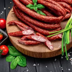 Kabanosy, z czego są? Z mięsa wieprzowego, przypraw i czosnku. Na desce z kabanosami pomidorki, szczypiorek i bazylia.
