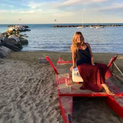 Donna sorridente su una barca rossa sulla spiaggia di Cattolica, Emilia Romagna, con il mare e i faraglioni sullo sfondo.