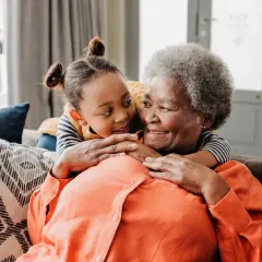 Ein süßer Moment zwischen Oma und Enkel. Das kleine Mädchen umarmt ihre Großmutter von hinten, beide lächeln sich liebevoll an.