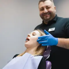 Uśmiechnięty dentysta w niebieskich rękawiczkach bada pacjentkę. Zespół Costena dba o zdrowie jamy ustnej.