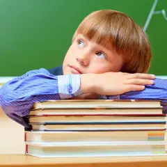 Chłopiec w niebieskiej koszuli opiera głowę na stosie książek, patrząc w górę.