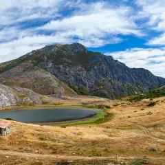 Pequeña cabaña de piedra junto a un lago ausente, rodeada de montañas escarpadas y prados secos bajo un cielo azul con nubes.