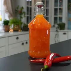 En flaska med hemmagjord sweet chili sås står på en mörk bänk, med två röda chilifrukter framför.