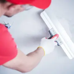 Ręce w białych rękawiczkach z czerwoną łopatką wygładzają tynk na ścianie.