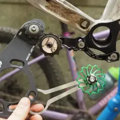 A hand holds a bike chain guide, part of a 1x drivetrain, near a bicycle frame. A green pulley wheel is also visible.