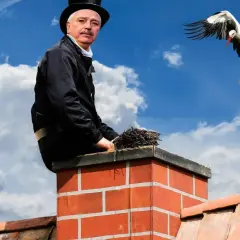 Kominiarz na dachu z miotłą, bocian w locie. PZU ubezpieczenie domu a przegląd komina to podstawa bezpieczeństwa.