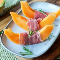 Melone mit Schinken, garniert mit Rosmarin, auf einem Teller. Ein Klassiker, der süße und salzige Aromen vereint.