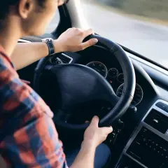 Uomo al volante di un'auto, con le mani sul volante. La sua camicia a quadri rossi e blu si abbina al cruscotto dell'eps auto.