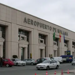 Lotnisko w Maladze, z charakterystycznym napisem na fasadzie. Przed budynkiem parkują samochody, a obok powiewają flagi.