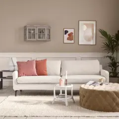 Beige ist die Farbe der Wände, des Sofas und des runden Hockers. Ein gemütliches Wohnzimmer mit weißen Dielen und einem Teppich.