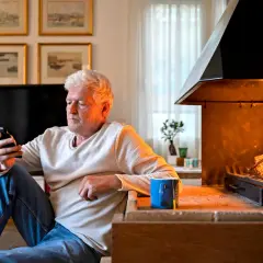 Mann sitzt vor modernem Kamin und schaut aufs Handy. Verschiedene Kamin Arten sind beliebt, dieser hier spendet wohlige Wärme.
