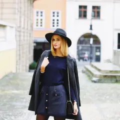 Elegancka kobieta w kapeluszu i płaszczu, w granatowym kolorze, pozuje na tle kamienic.