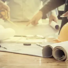 Zespół analizuje plany, dyskutując, ile trwa podział działki. Na stole leżą zrolowane projekty, miarka i żółty kask.