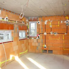 W trakcie budowy domu, widoczne są **domowe instalacje elektryczne** na ścianach z cegły, z przewodami i puszkami.