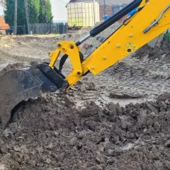 Żółta koparka marki JCB pracuje na budowie, kopiąc ziemię. Widać jej ramię i łyżkę zagłębiającą się w błotniste podłoże.