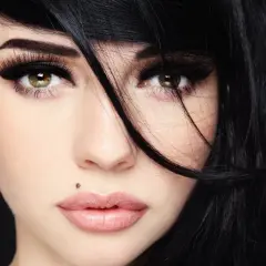 Kobieta z czarnymi włosami i makijażem podkreślającym oczy. Widoczny monroe piercing nad górną wargą.