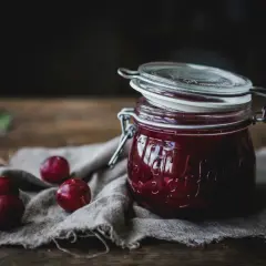 En burk med hemlagad plommonmarmelad med kanel, omgiven av färska plommon på en linneduk.
