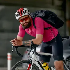 Mężczyzna w różowej koszulce i czarnych spodenkach, kasku i okularach rowerowych, z plecakiem, pokazuje, jak się ubrać na rower.