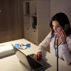 Młoda kobieta, pracująca jako dyspozytor, odbiera telefon przy biurku z laptopem i dokumentami.