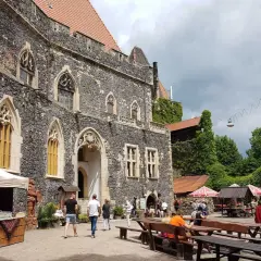 Kamienny grodziec z gotyckimi oknami, gdzie turyści spacerują i odpoczywają przy drewnianych ławach.