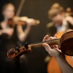 Dłoń trzymająca gryf skrzypiec, w tle dwie kobiety grające na instrumentach smyczkowych. To preludium do pięknej muzyki.