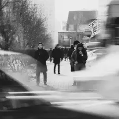 Ludzie na chodniku, rozmyci przez długi **shutter speed**. W tle drzewa i budynki.