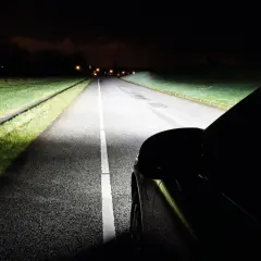 Jasny żarnik reflektora samochodu oświetla nocną drogę, ukazując trawę i zarys asfaltu.