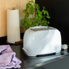 Ein weißer Toaster steht auf einer schwarzen Arbeitsplatte, bereit für die Reinigung. Daneben liegen ein lila Tuch und eine Küchenrolle.