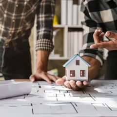 Dwóch mężczyzn analizuje projekt budowlany domu. Na stole leżą plany, makieta domu i kask.
