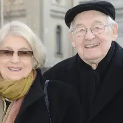 Uśmiechnięta para: Andrzej Wajda i jego żona, w eleganckich płaszczach i szalikach, pozują do zdjęcia na tle architektury.
