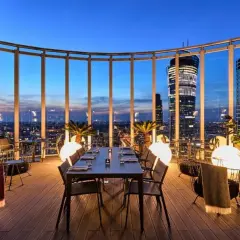 Przytulna restauracja Warszawa na dachu z widokiem na nocne miasto. Stoły i krzesła, palmy i kule świetlne tworzą magiczną atmosferę.
