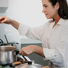 Kobieta w białej koszuli gotuje, nabiera drewnianą łyżką sos z garnka. Pokazuje, jak wyciągnąć tłuszcz z sosu, by był lżejszy.