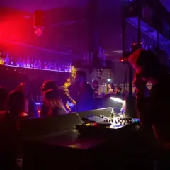 Un hot night club con luci rosse e viola, gente che balla e un DJ al lavoro. L'atmosfera è vibrante e piena di energia.