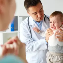 Lekarz uspokaja płaczące dziecko, którego rodzice sprawdzają skoki rozwojowe w tabeli.