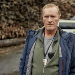 En man med intensiv blick, klädd i jacka och väst, bär en radio och ett id-kort. Bakom honom syns en polisbil och en vedhög. En scen från en norsk serie.