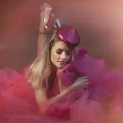 Urocza sesja glamour: blondynka w różowej tiulowej sukni i błyszczącej beretce, z makijażem oczu w tym samym kolorze, leży na ziemi.