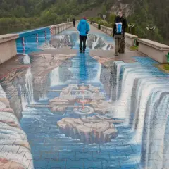 Niesamowita iluzja 3D na zapora w Niedzicy, gdzie wodospady i kamienie zdają się wypływać z chodnika. Dwie osoby spacerują po tej artystycznej instalacji.